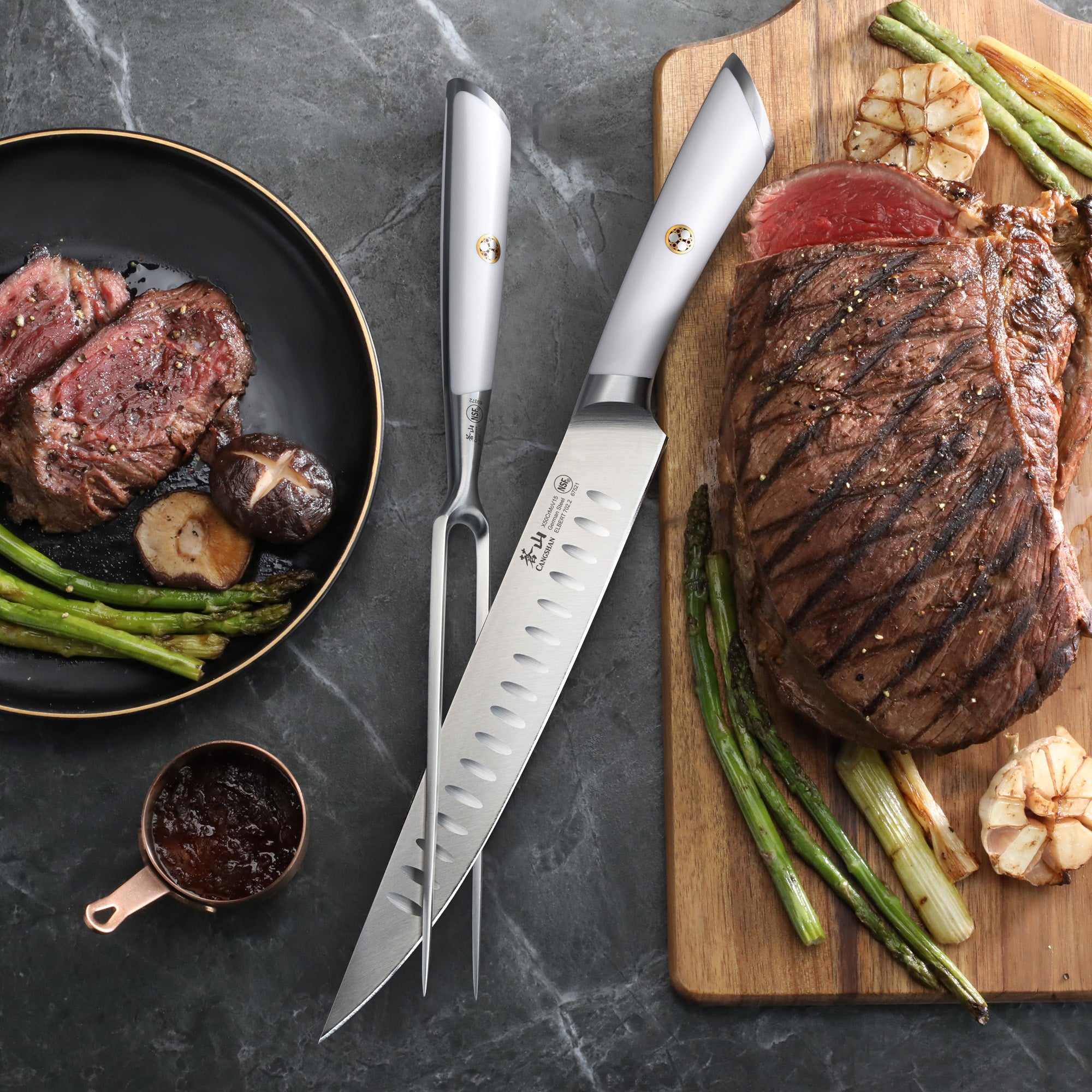 Two-piece carving set with a carving knife and fork next to grilled steak, asparagus, and garlic on a cutting board and plate