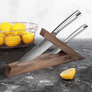 
                    
                      Two stainless steel knives stored in a triangular walnut wood knife block on a dark countertop with a halved lemon nearby.
                    
                  