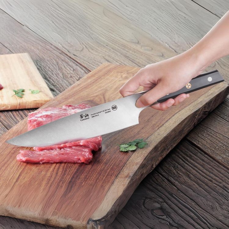 Hand holding a chef knife slicing raw meat on a wooden cutting board with herbs nearby.