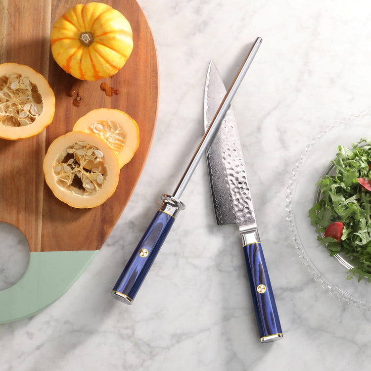Blue-handled chef's knife and honing steel on marble surface next to wooden board with sliced squash and a bowl of greens.