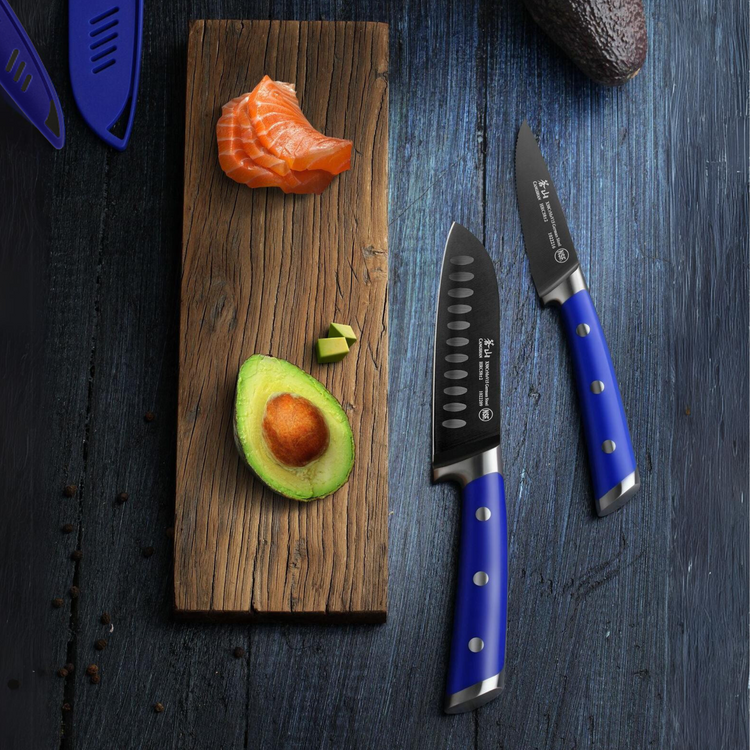 Half an avocado with two cubes, a piece of sliced salmon on a wooden board, and two blue-handled knives on a dark surface.