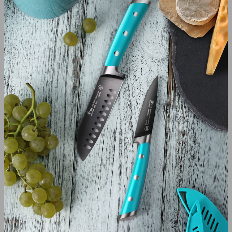Two turquoise-handled knives on a wooden surface, next to grapes and cheese on a black slate board.