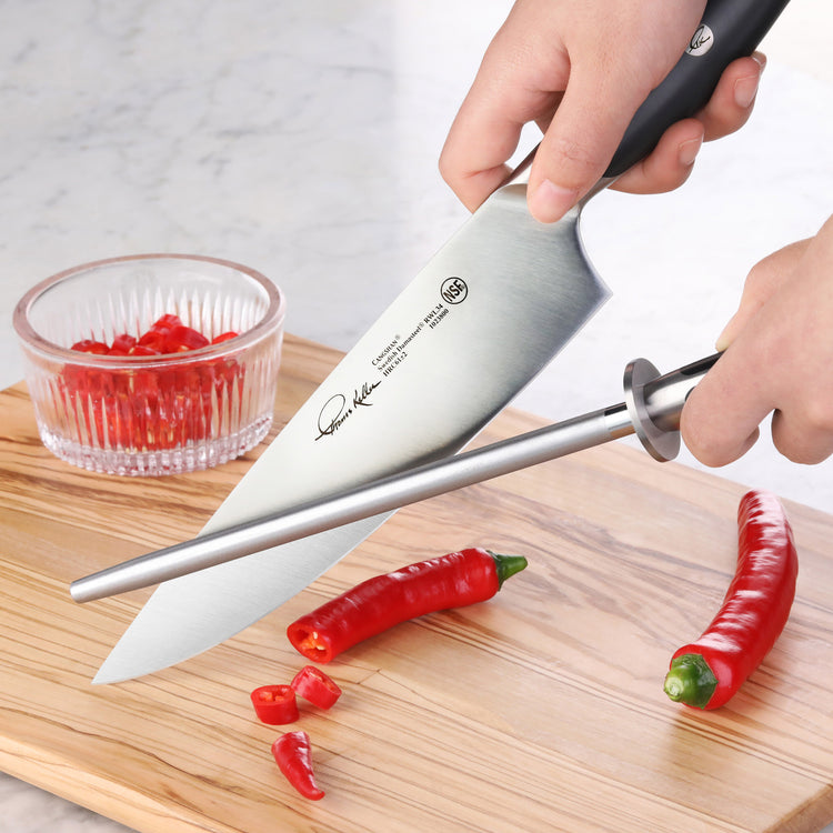 Person sharpening a chef's knife with a honing steel on a wooden cutting board next to red chili peppers.