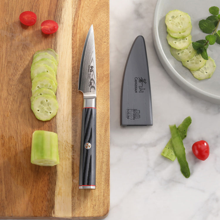 3.5-inch paring knife with black handle and sheath beside sliced cucumber and cherry tomatoes on wood and marble surfaces.