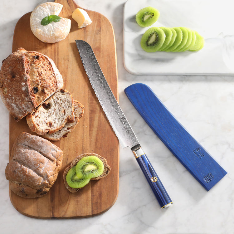 Bread knife with blue handle and sheath next to sliced bread, cheese, and sliced kiwi on wooden and marble boards.
