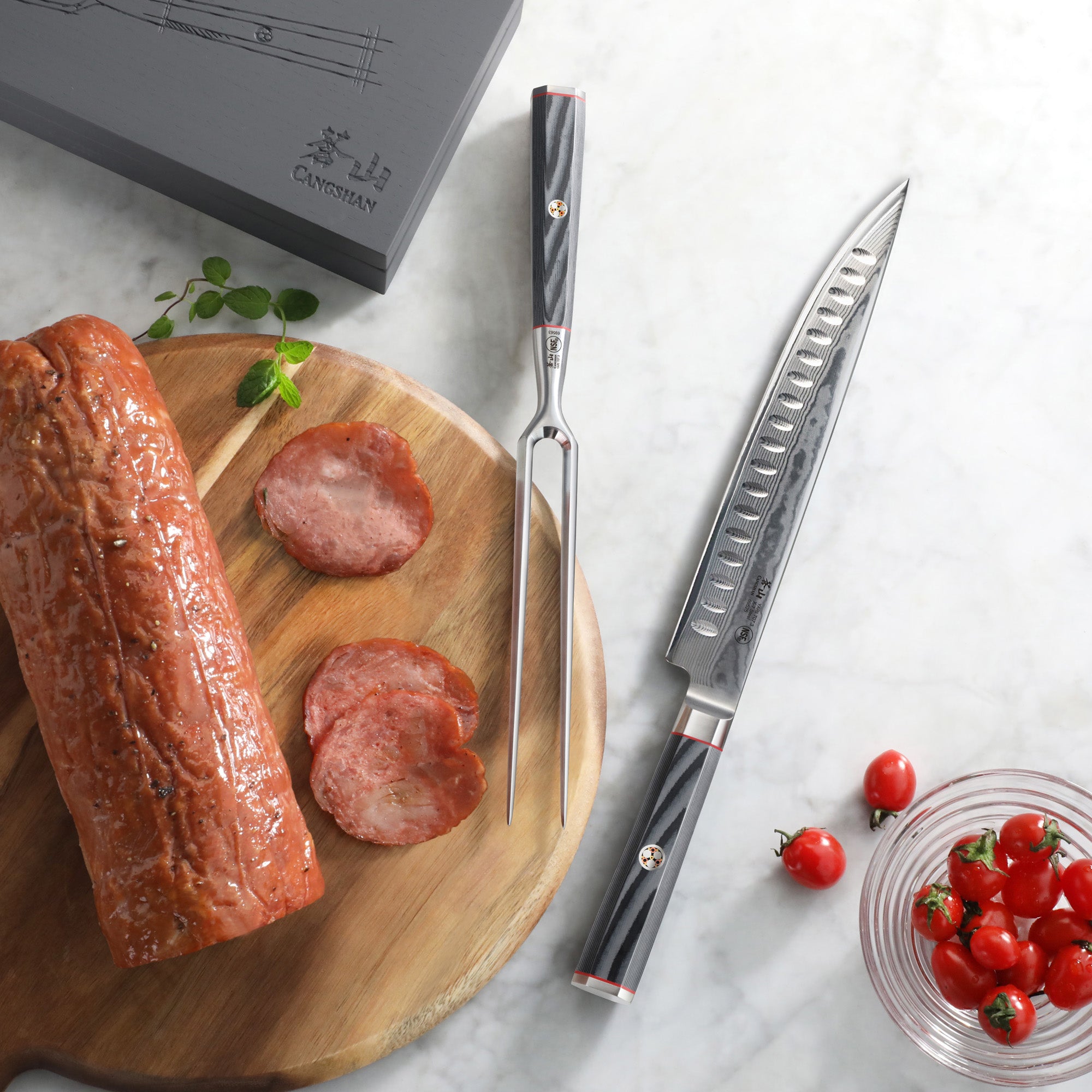 Carving knife and fork with black handles on marble surface, sliced meat on wooden board, cherry tomatoes in glass bowl.