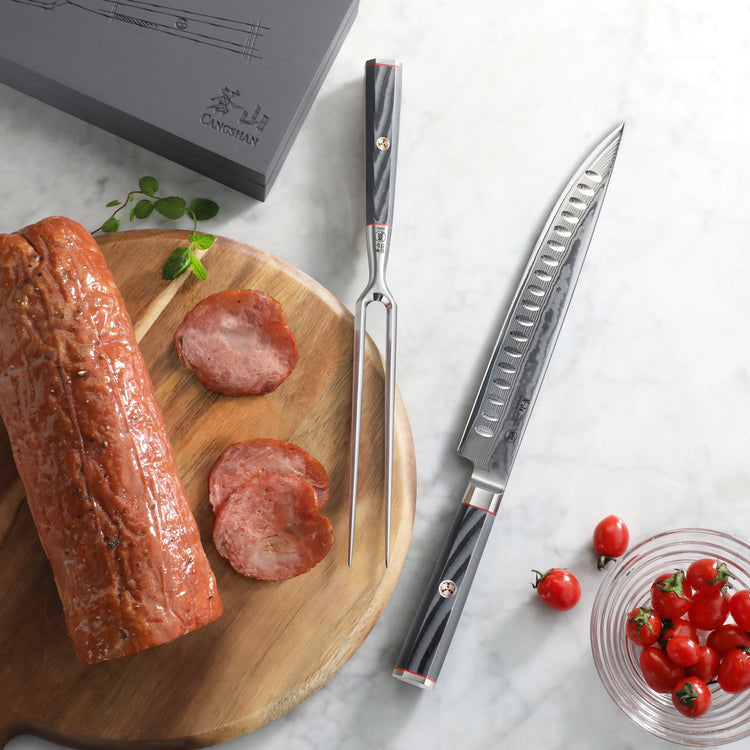 Carving knife and fork with black handles on marble surface, sliced meat on wooden board, cherry tomatoes in glass bowl.