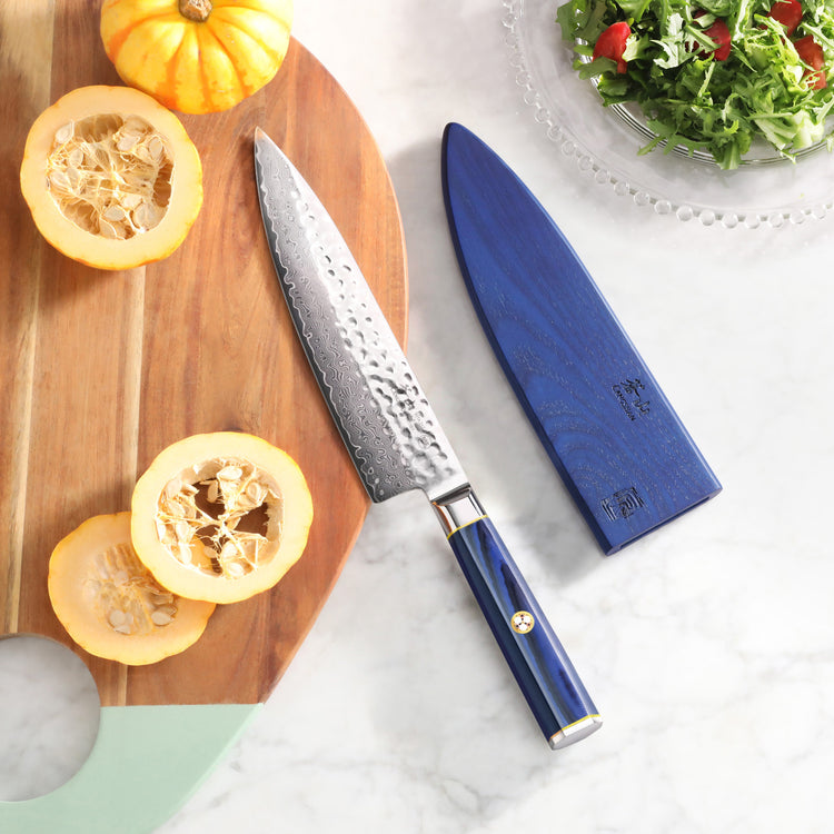 8-inch chef knife with hammered Damascus steel blade and blue sheath on marble surface next to cut squash and salad.