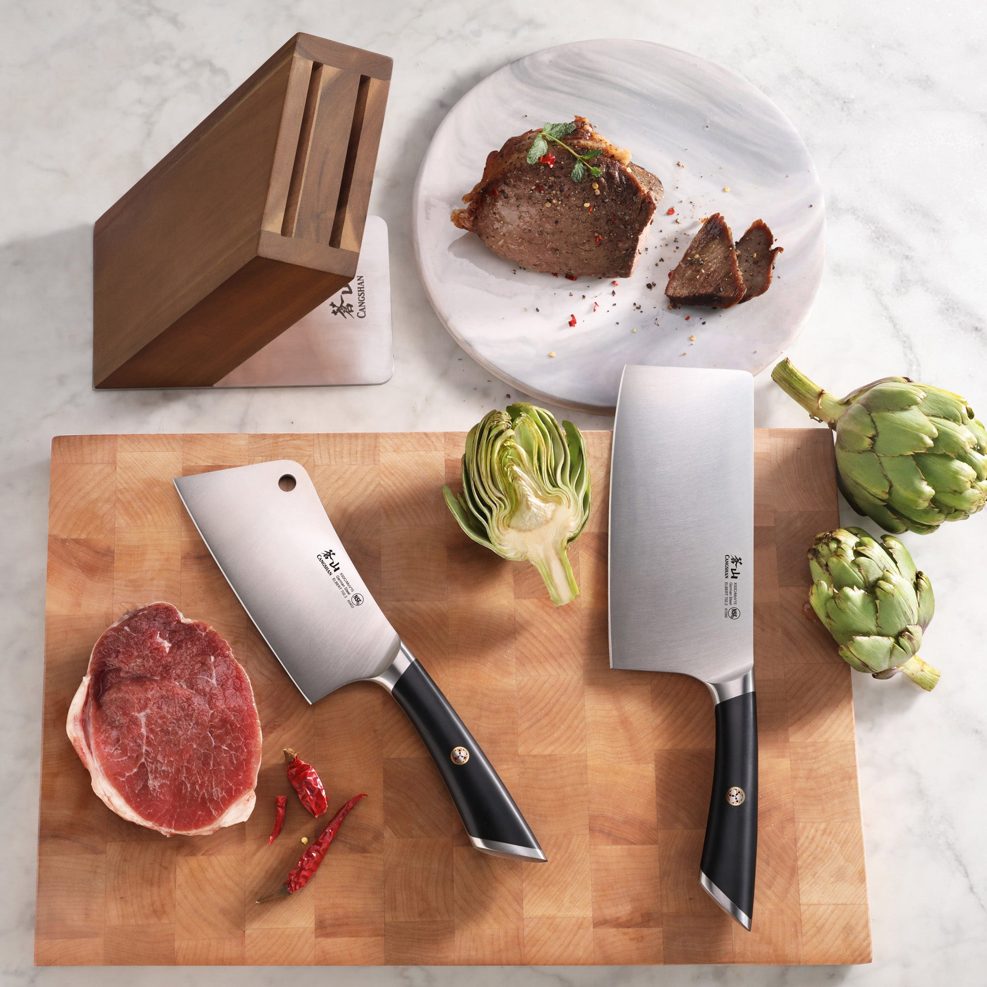 Two cleaver knives with black handles on a wooden cutting board with raw meat, artichokes, chili peppers, and a wooden knife