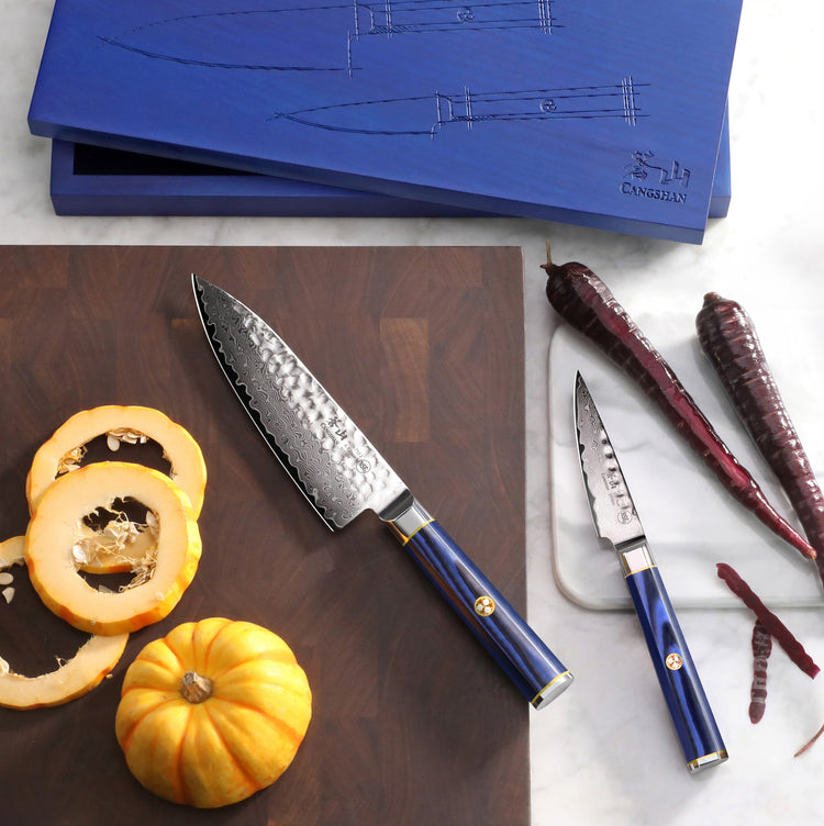 Two blue-handled knives with hammered blades on a cutting board, surrounded by sliced squash, whole squash, and red chili pep