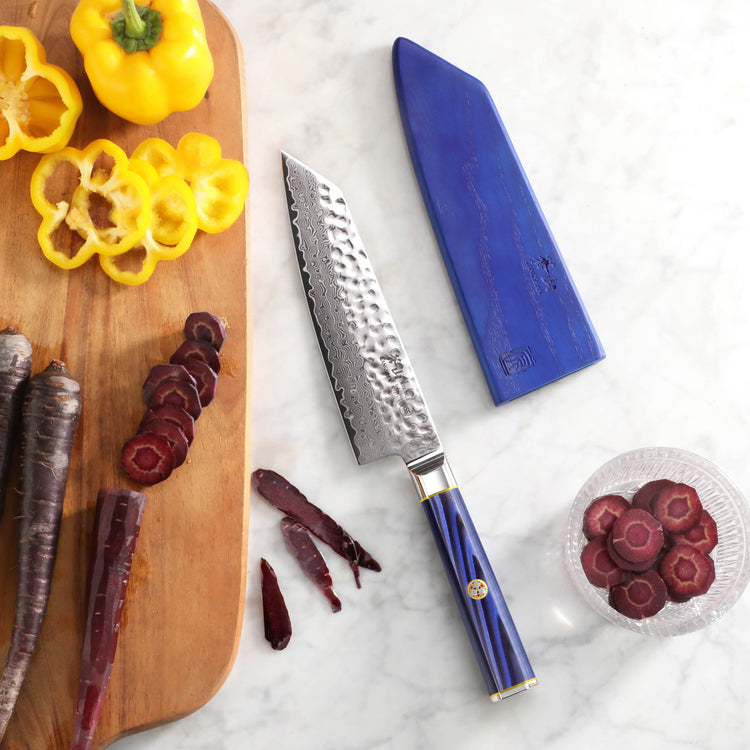 7-inch Kiritsuke knife with hammered blade and blue handle, next to blue sheath, sliced yellow bell pepper and purple carrots