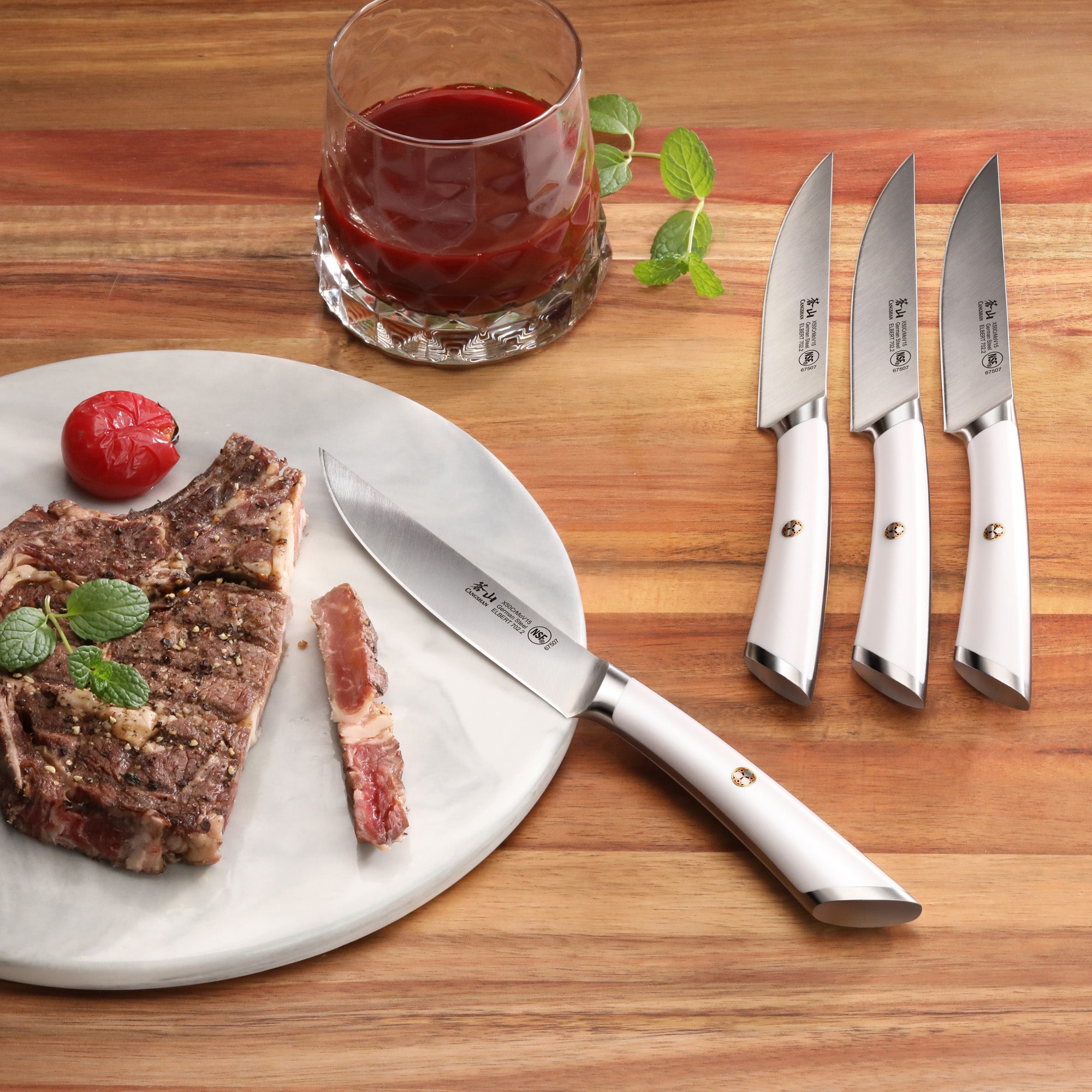Four steak knives with white handles on a wooden surface, one cutting a cooked steak on a white plate next to a glass of red