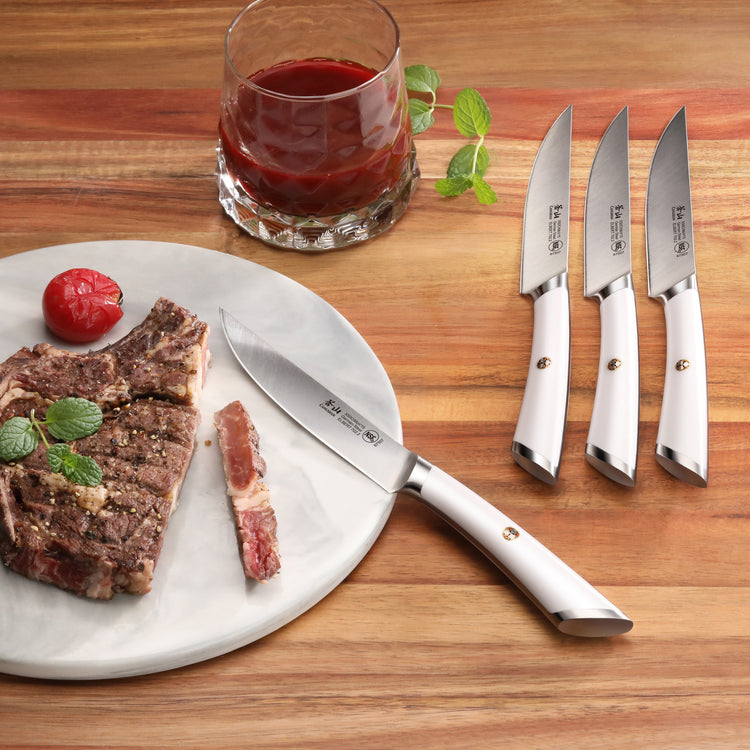 Four steak knives with white handles on a wooden surface, one cutting a cooked steak on a white plate next to a glass of red