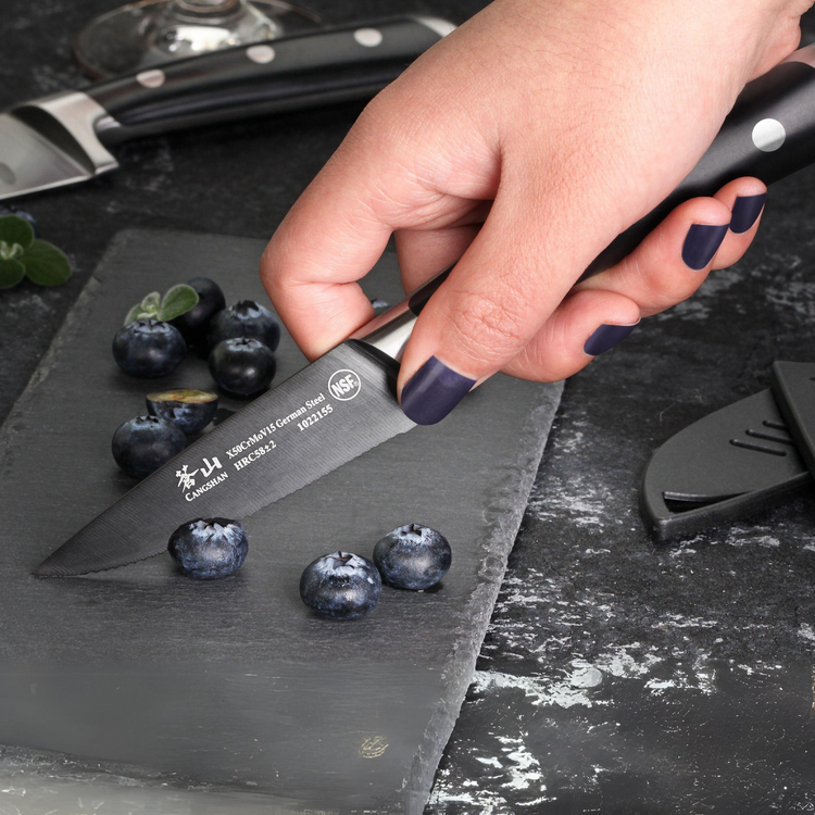 Hand with dark nail polish holding a small serrated knife cutting blueberries on a dark slate board.