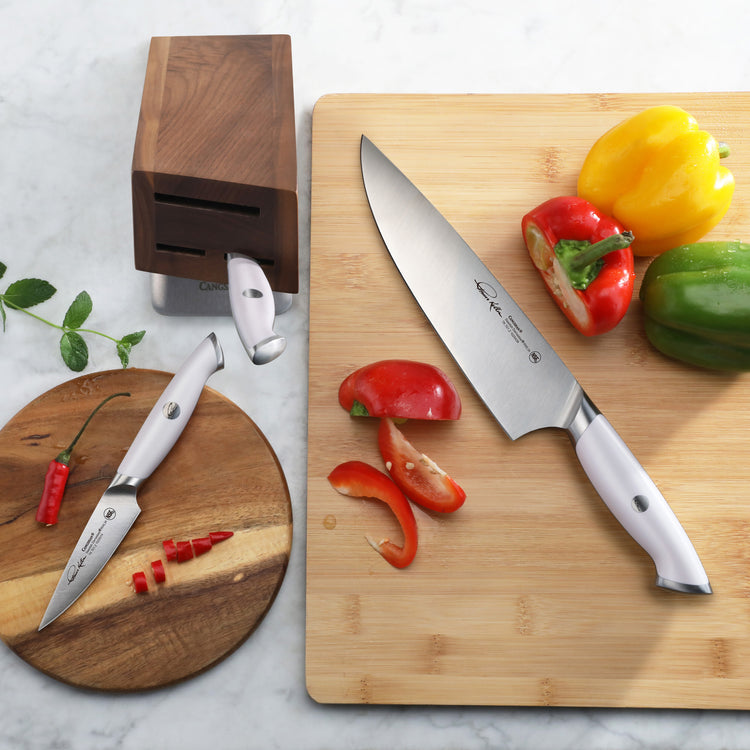 Two stainless steel knives with white handles, one slicing a red bell pepper on a wooden cutting board, next to a wooden knif