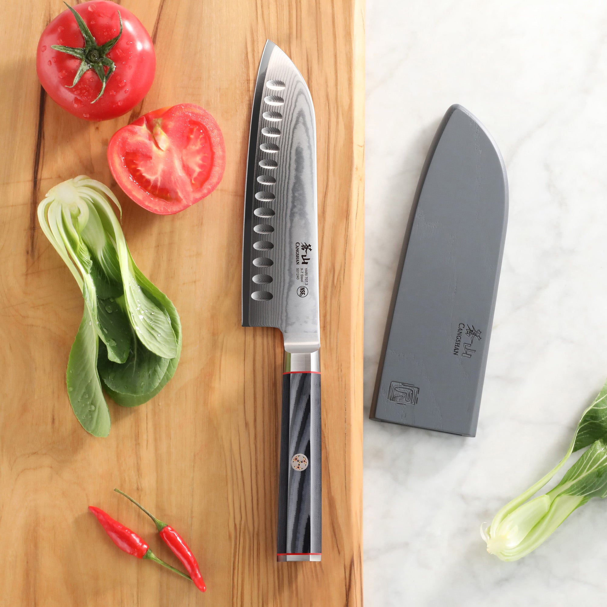7-inch Santoku knife with patterned blade and black handle on wooden board with tomatoes, chili peppers, and sheath nearby.