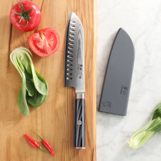 
                    
                      7-inch Santoku knife with patterned blade and black handle on wooden board with tomatoes, chili peppers, and sheath nearby.
                    
                  