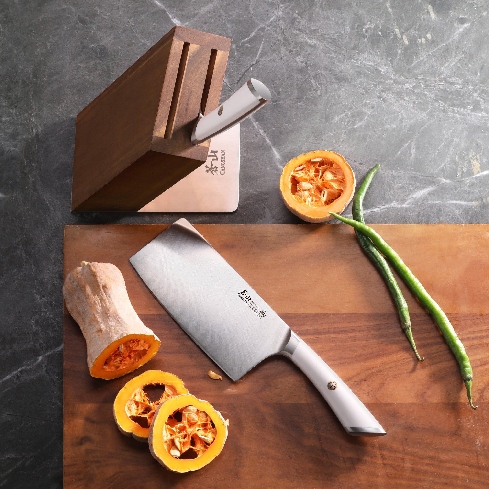 Cleaver knife on wooden board with sliced squash, green chili peppers, and a wooden knife block on gray surface.