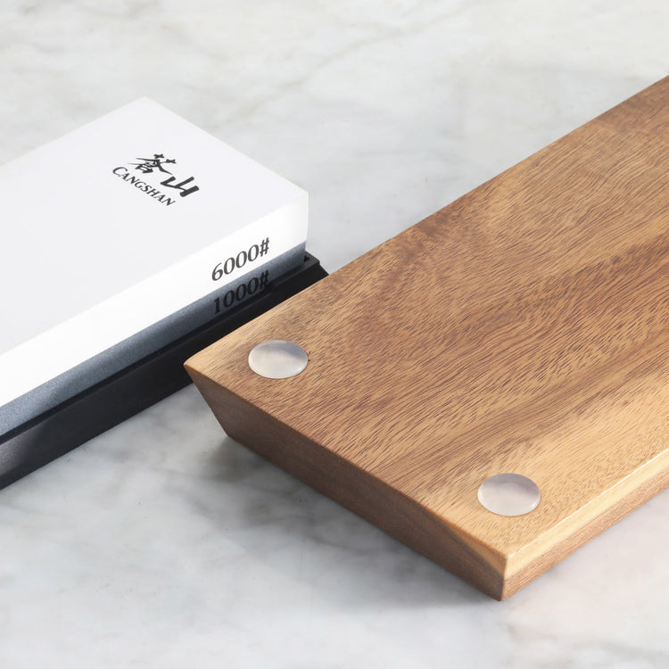Dual-sided sharpening stone with 1000 and 6000 grit resting on a black base next to a wooden knife handle.
