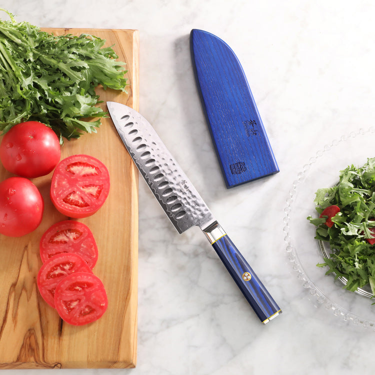 Santoku knife with blue handle and sheath next to sliced tomatoes and leafy greens on a marble surface.