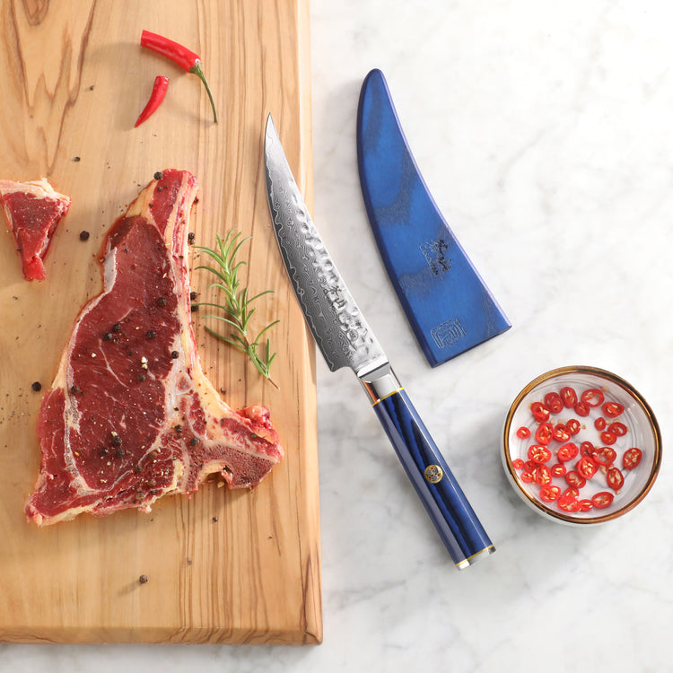 6-inch boning knife with blue handle and sheath next to raw steak, rosemary, chili peppers, and sliced chili in bowl.