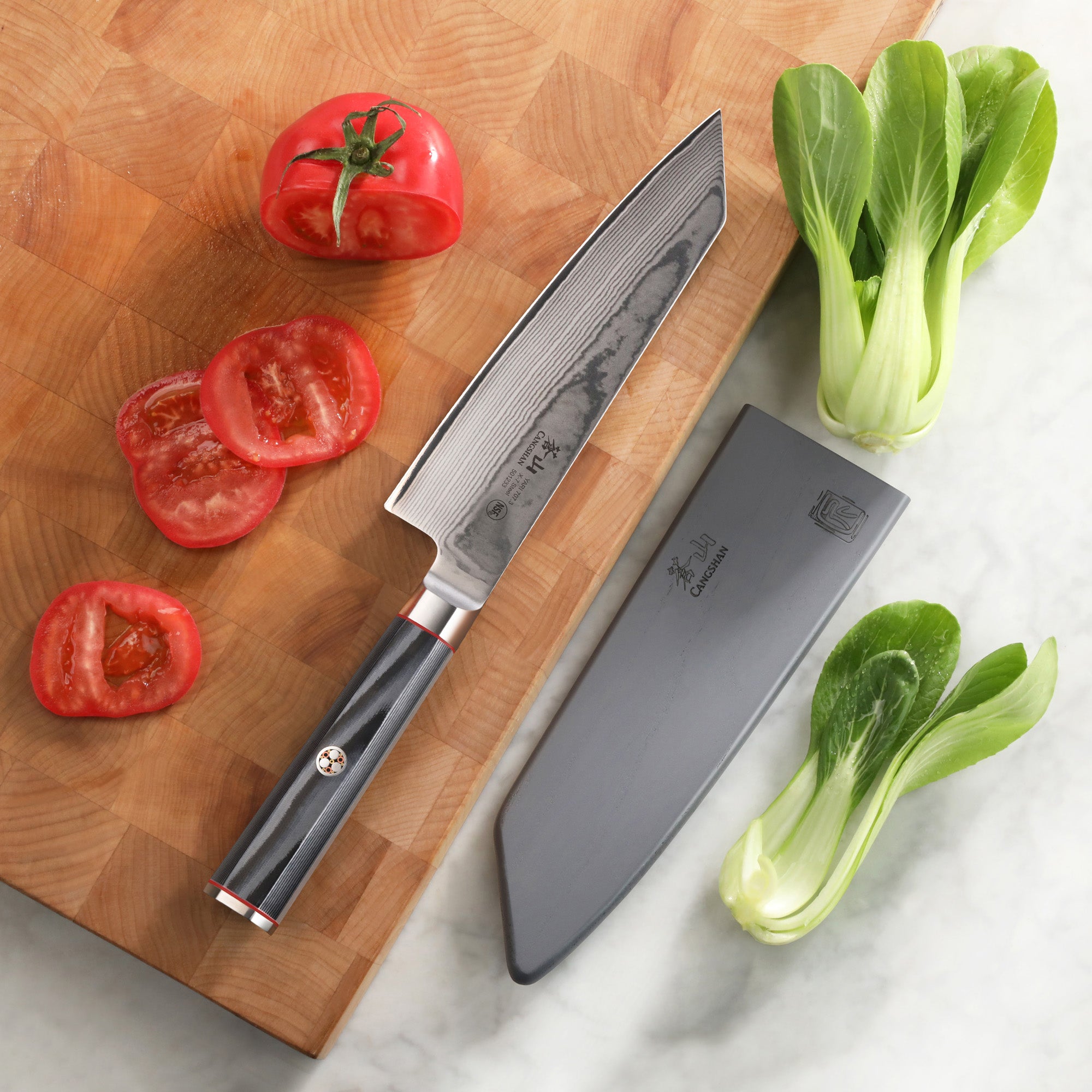 7-inch Kiritsuke knife with patterned blade and black handle, sheath, sliced tomato, and bok choy on cutting board and marble