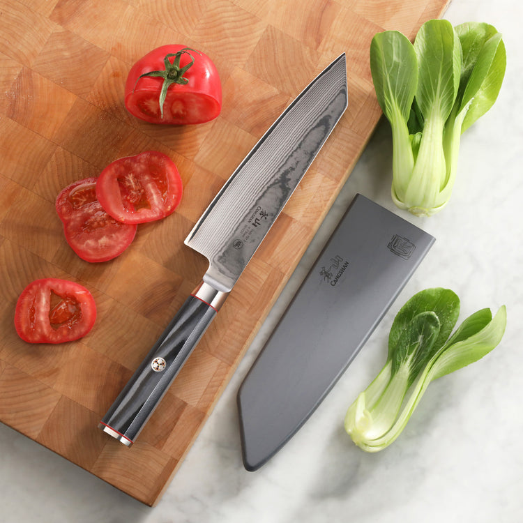 7-inch Kiritsuke knife with patterned blade and black handle, sheath, sliced tomato, and bok choy on cutting board and marble