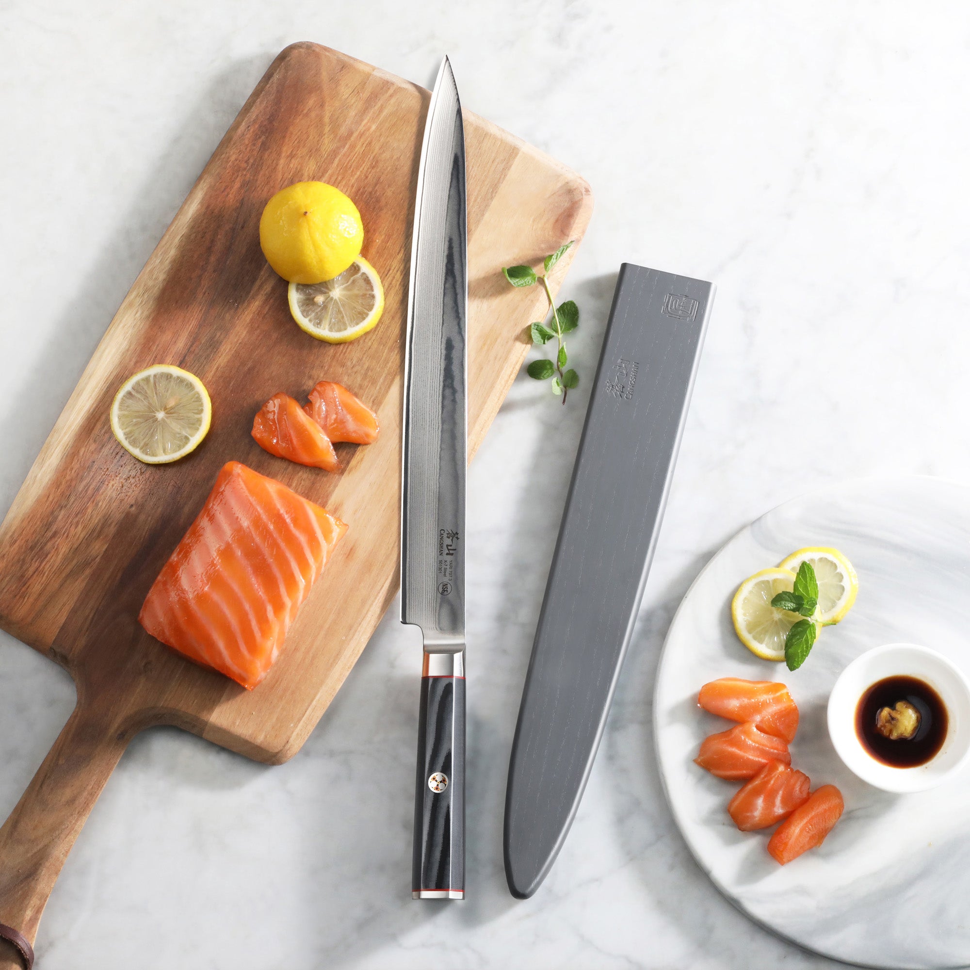12-inch sashimi knife with black handle and sheath beside salmon slices, lemon, and soy sauce on wooden and marble surfaces