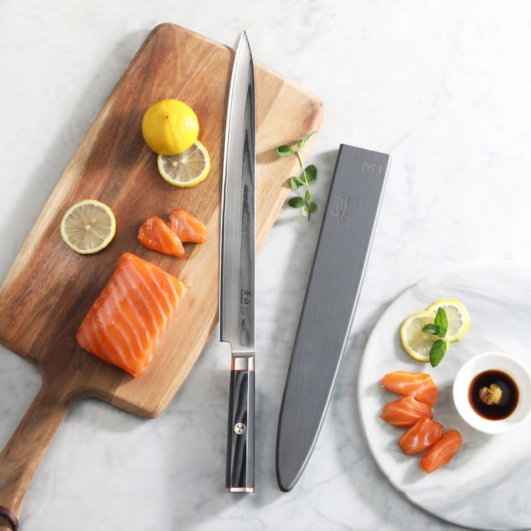 12-inch sashimi knife with black handle and sheath beside salmon slices, lemon, and soy sauce on wooden and marble surfaces