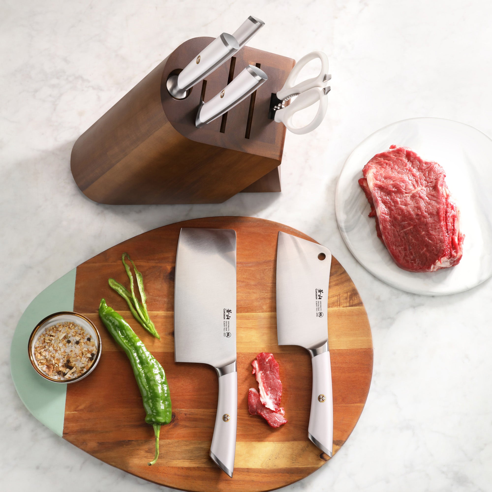 Wooden knife block with three knives and scissors, two cleaver knives on a cutting board with green peppers and raw meat.