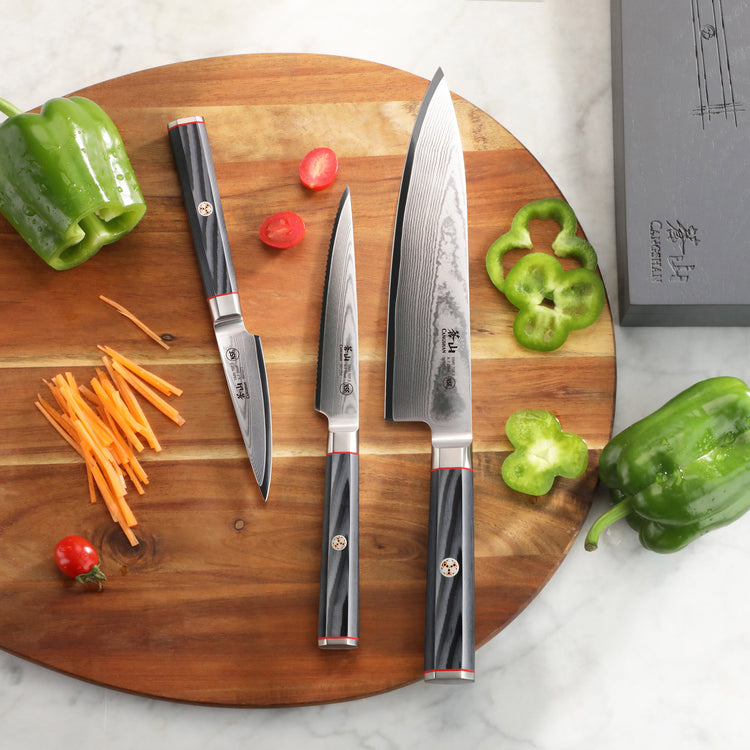 Three knives with black handles and Damascus steel blades on a wooden cutting board with sliced vegetables and a gray box nea
