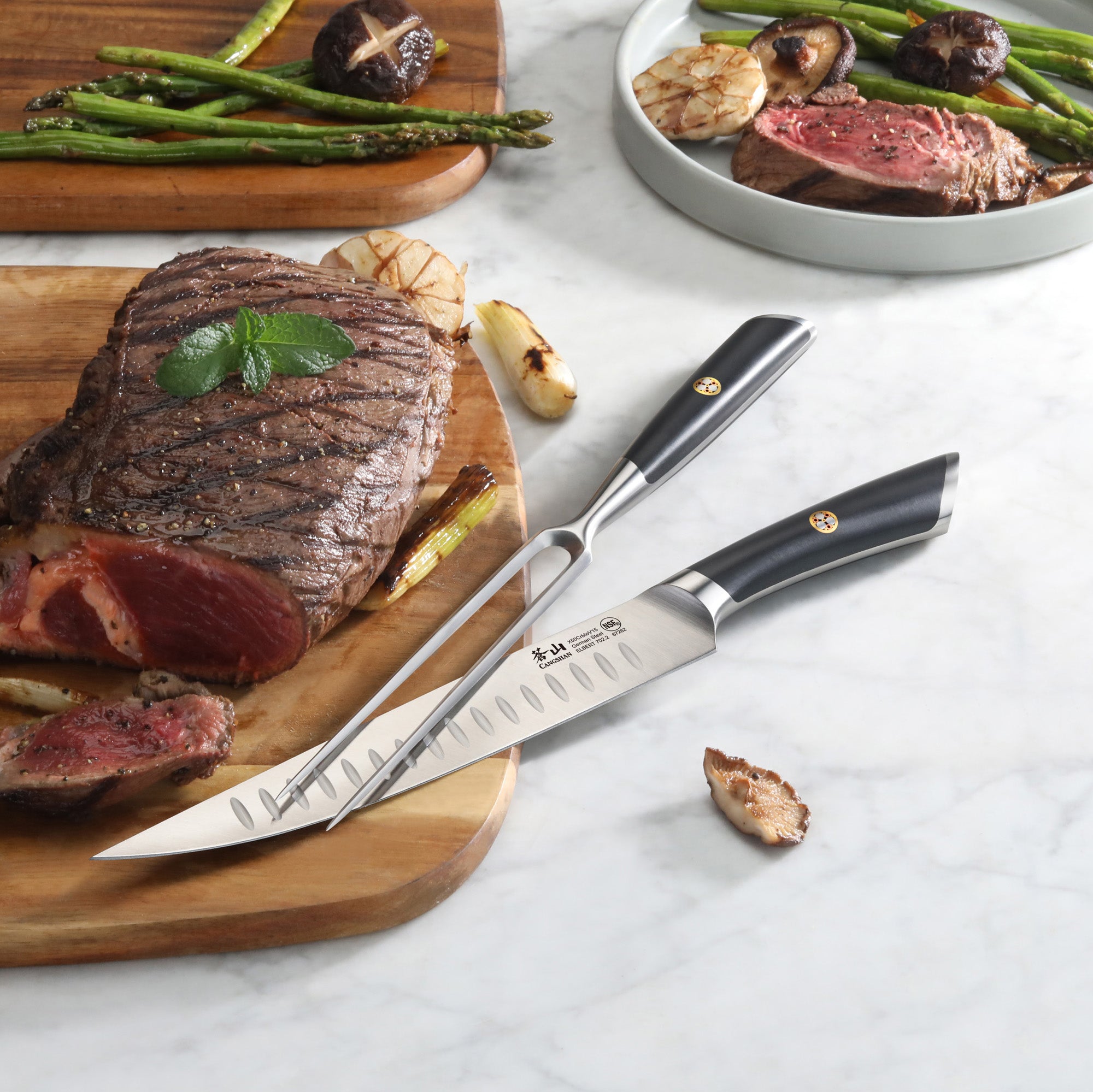 Two-piece carving set with a carving knife and fork beside sliced grilled steak and vegetables on a marble surface.