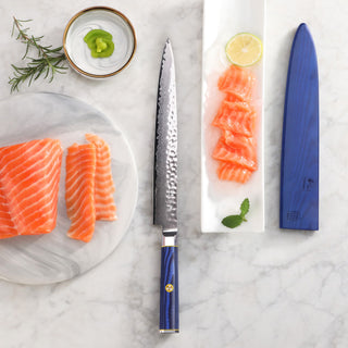 
                    
                      10-inch sashimi knife with blue handle and sheath, next to sliced salmon, lemon, wasabi, and herbs on marble surface.
                    
                  