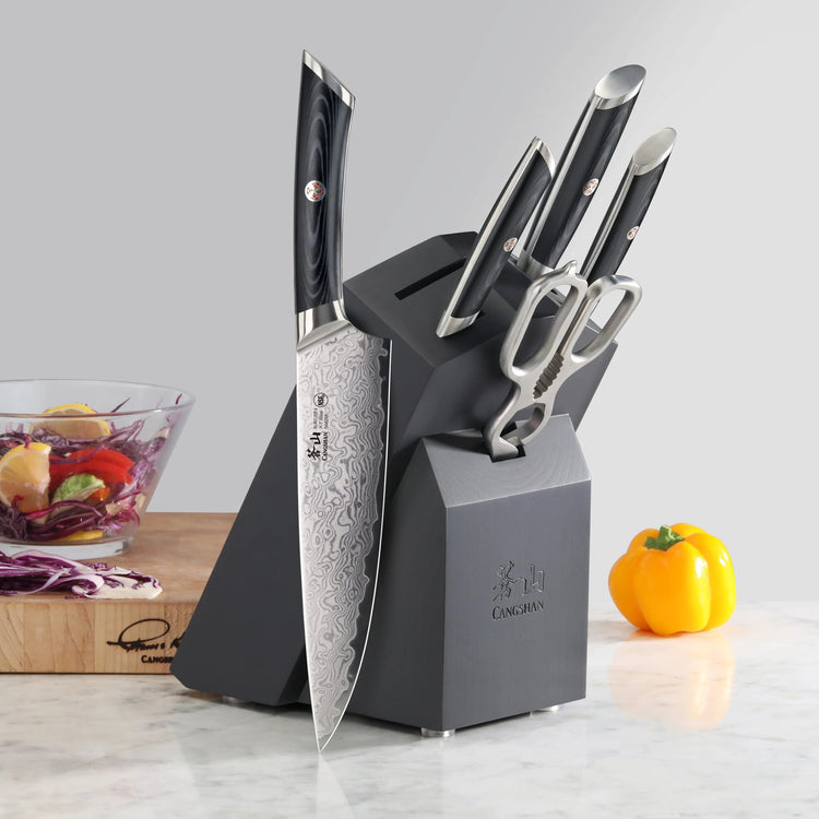 Six-piece knife set with patterned blades and black handles in a black block, next to a yellow bell pepper and a salad bowl.
