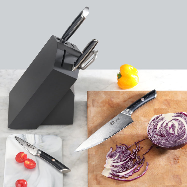 Black knife block with three knives inserted, one knife on a cutting board slicing purple cabbage, cherry tomatoes on a marbl