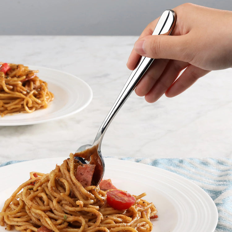 Hand holding a polished stainless steel serving fork lifting spaghetti with tomato and meat from a white plate.
