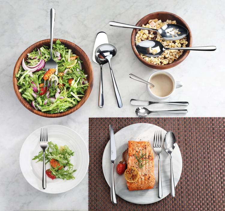 Serving spoon with mirror finish placed in wooden bowls with salad and cereal, alongside plated salad and salmon meal with fl