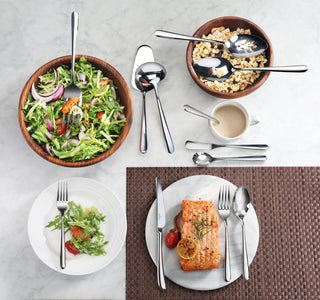 
                    
                      Two stainless steel spoons with mirror finish placed in wooden bowls with salad and cereal, alongside a table setting with sa
                    
                  