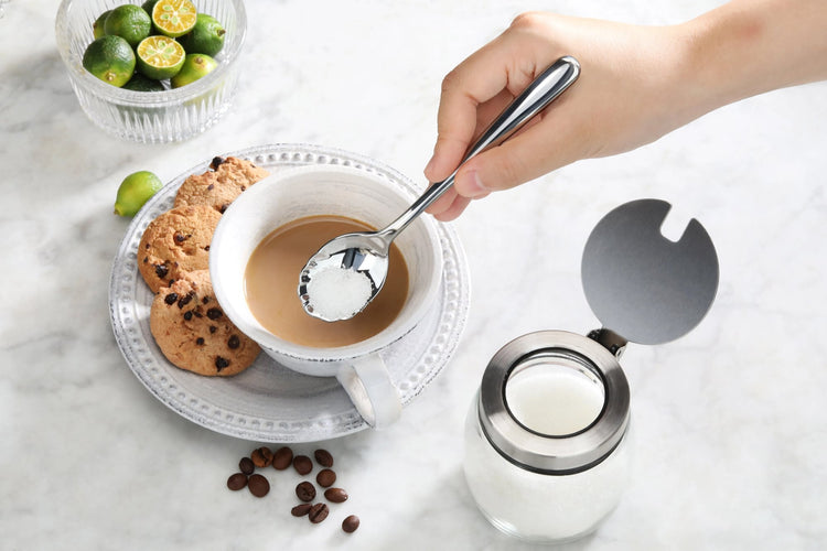 Hand holding a polished stainless steel sugar spoon over a cup of coffee with cookies and a sugar container nearby.