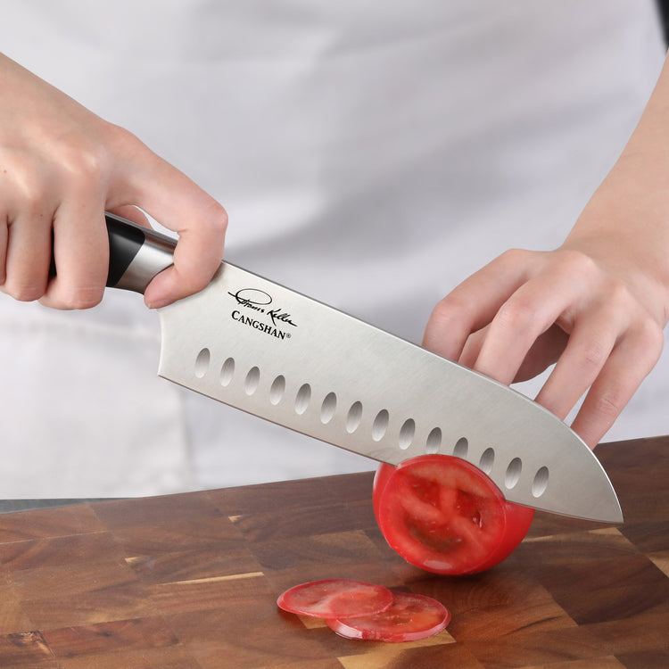 Hand holding a Santoku knife slicing a tomato on a wooden cutting board.