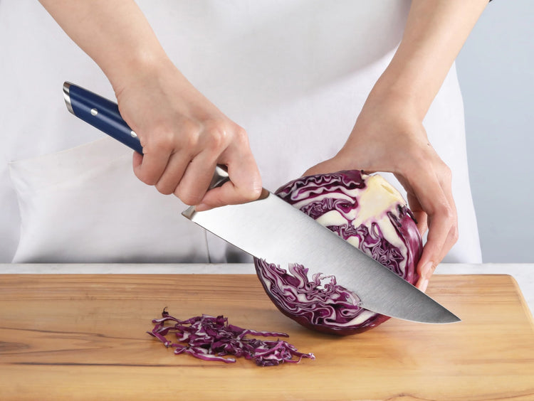 Hands slicing a halved purple cabbage on a wooden cutting board with a large knife featuring a blue handle.
