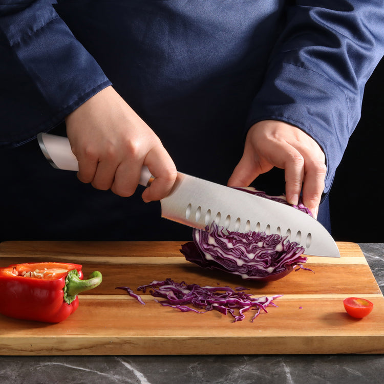 Hands slicing purple cabbage on a wooden cutting board with a white-handled Santoku knife, next to a red bell pepper and cher