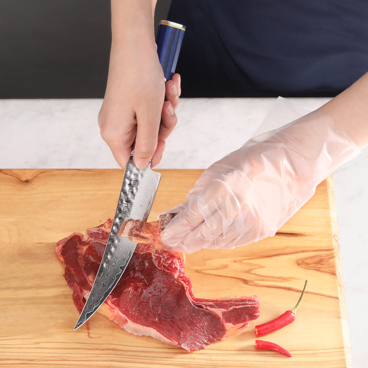 Hand wearing a plastic glove holding a blue-handled boning knife cutting raw meat on a wooden board with two red chili pepper