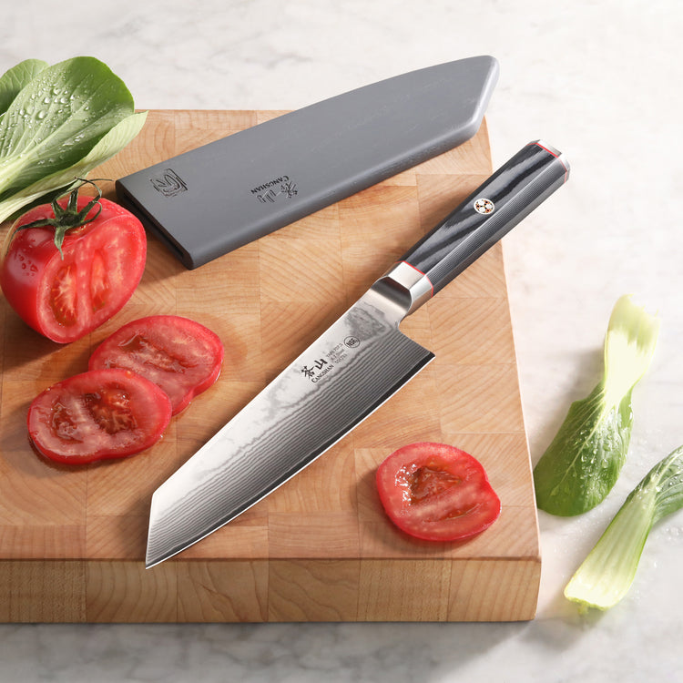 7-inch Kiritsuke knife with Damascus steel blade and black handle on wooden cutting board with sliced tomato and sheath nearb