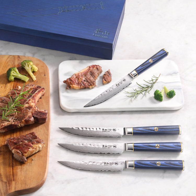 Four steak knives with blue handles and Damascus steel blades displayed with cooked steak, broccoli, and a blue box.