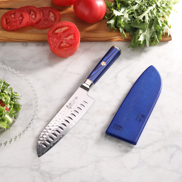 Santoku knife with blue handle and matching sheath on marble surface, next to sliced tomatoes and greens.