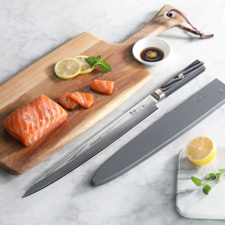 12-inch sashimi knife with sheath beside sliced salmon on wooden board with lemon, soy sauce, and herbs.