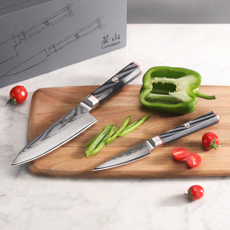 Two Damascus steel knives with black handles on a wooden cutting board, alongside sliced green bell pepper and cherry tomatoe