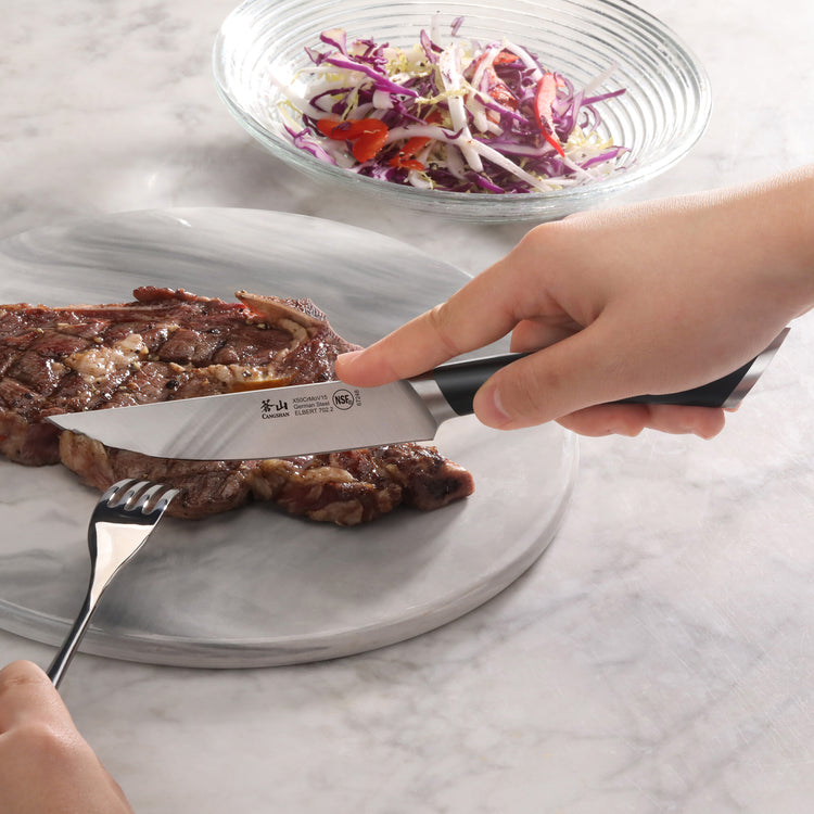 Hand holding a steak knife cutting a grilled steak on a plate with a fork, salad bowl in the background.