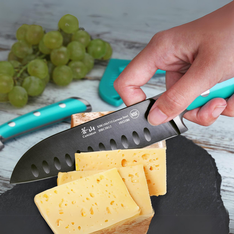 Hand holding a turquoise-handled knife slicing semi-hard cheese on a slate board with green grapes in the background.